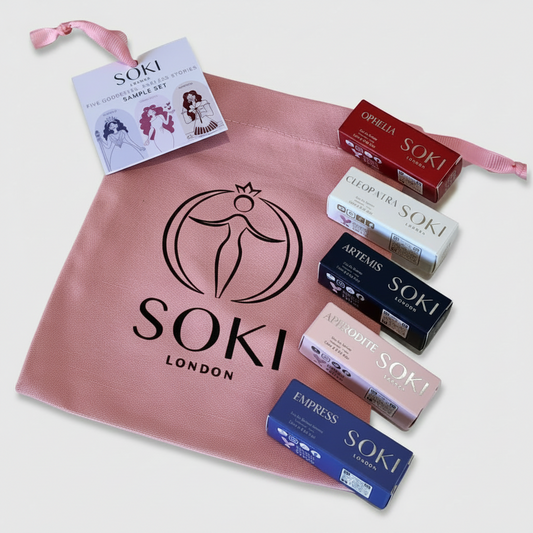 Soki London fine fragrance sample set featuring a dusty pink drawstring pouch with the Soki logo, five individual cosmetic boxes in red, white, black, pink, and blue, and a sample card, arranged on a light gray background.