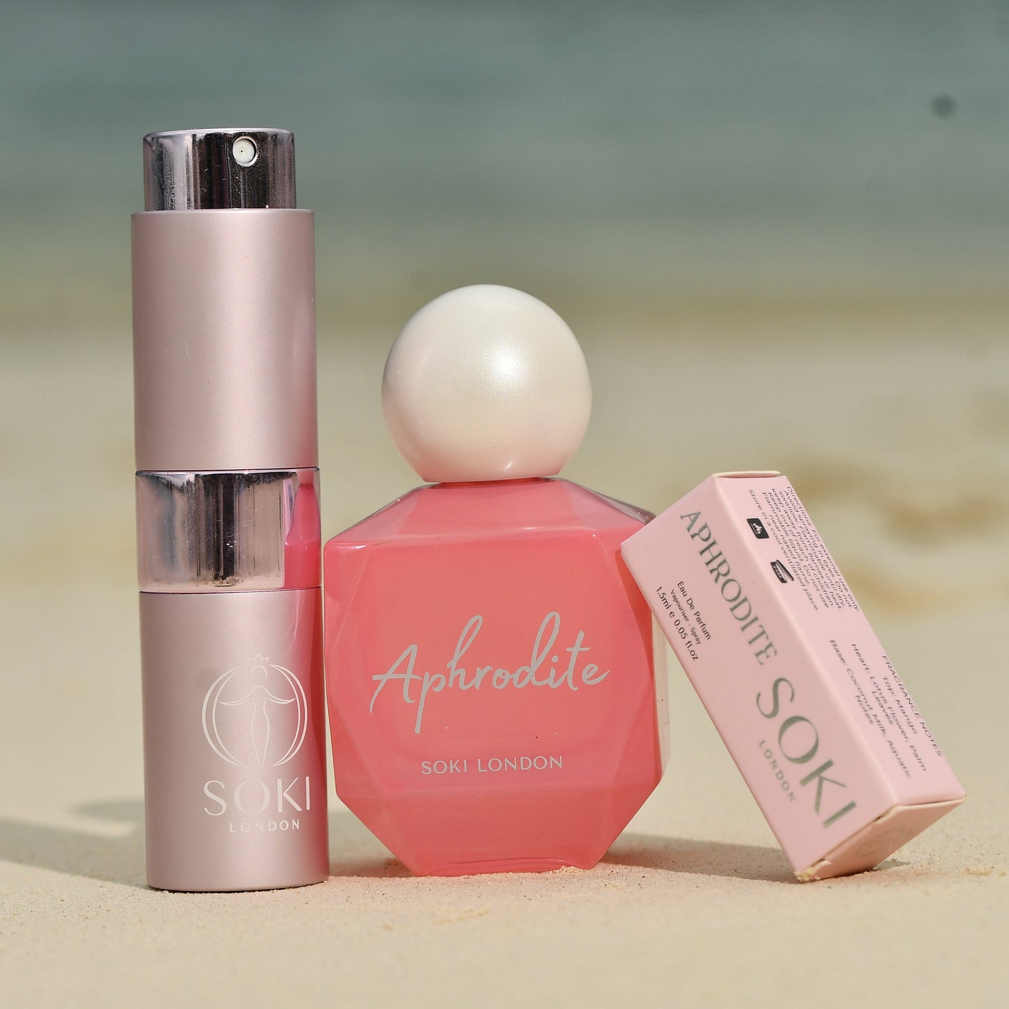 Soki Aphrodite perfume and matching skincare set on a sandy beach background. The set includes a pink hexagonal Aphrodite perfume bottle with a white pearl cap, a metallic pink Soki bottle, and a pink Aphrodite box.