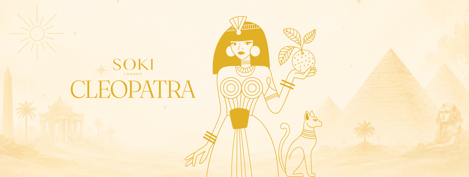 Egyptian-themed graphic banner with the text 'SOKI LONDONCLEOPATRA' in gold lettering, featuring a stylized gold illustration of Cleopatra holding an apple, a cat, pyramids, and a sun on a pale cream background.